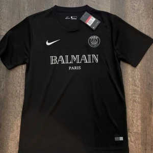 Svart PSG x Balmain fotbollströja Nike - Snygg svart fotbollströja från Nike i samarbete med PSG och Balmain. Tröjan har korta ärmar, rund hals och tydliga vita loggor framtill. Tillverkad i lätt och funktionellt material som passar perfekt för träning eller match. fraktar inom 24 timmar.