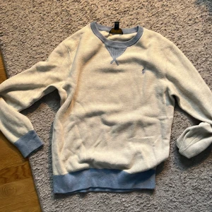 Vit och ljusblå tröja Polo Ralph Lauren - Säljer en stilren vit tröja från Polo Ralph Lauren med ljusblå ribbade muddar och krage. Tröjan har lång ärm och klassisk broderad logga på bröstet. Perfekt för dig som gillar en clean och avslappnad look.