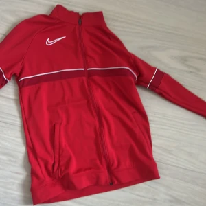 Röd Nike träningsjacka XL barn - Röd träningsjacka från Nike i storlek XL med hel dragkedja, vita detaljer och klassisk Swoosh-logga på bröstet. Jackan har ståkrage, fickor framtill och är gjord i Dri-FIT polyester som andas. Perfekt för sport eller chill.