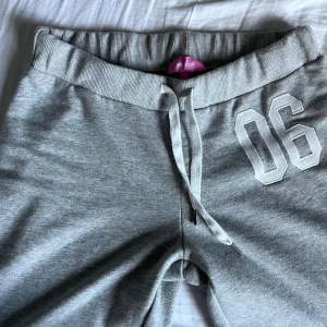 Grå mjukisbyxor från Edikted - Gråa mjukisbyxor från Edikted  med bred resår i midjan och vit dragsko. Snygg detalj med siffran 06 tryckt på ena benet. Byxorna har raka ben och är gjorda i mjukt bomullsmaterial, perfekta för chill dagar eller träning. Nypris: 829
