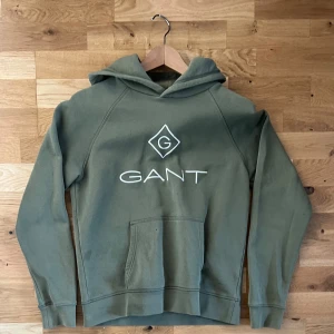 Olivgrön hoodie från GANT - Olivgrön hoodie från GANT med broderad logga i vitt framtill. Klassisk känguruficka och huva. Skön passform och mjukt material, perfekt för chill dagar. Snygg och enkel stil som funkar året runt.