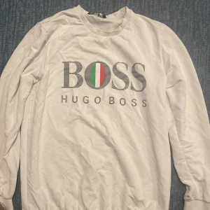 Vit sweatshirt från Hugo Boss - Vit sweatshirt från Hugo Boss med stort BOSS-tryck och logga framtill. Tröjan har rund halsringning och långa ärmar. Snygg detalj med röd och grön accent i loggan. Perfekt för en clean och stilren look.