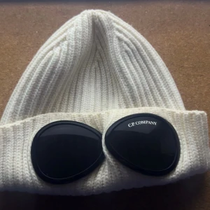 Vit C.P. Company Goggle Beanie - Ribbstickad vit mössa från C.P. Company med ikoniska svarta goggle-linser på uppviket. Snygg och unik design som sticker ut, perfekt för dig som gillar streetwear och vill ha något extra. Materialet är mjukt och stickat för skön komfort.