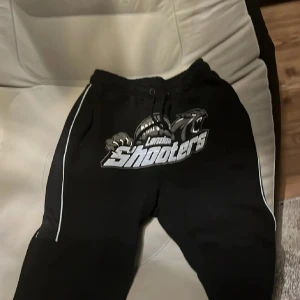 Svarta London Shooters mjukisbyxor - Svarta mjukisbyxor med stort London Shooters-tryck framtill och vita ränder längs benen. Byxorna har snörning i midjan och är perfekta för en avslappnad stil. Mjukt material och sportig look.