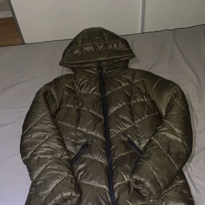 Olivgrön pufferjacka från Amisu - Säljer en olivgrön pufferjacka från Amisu med huva och dragkedja framtill. Jackan har quiltad design och två snedställda fickor med dragkedja. Perfekt för kalla dagar och riktigt snygg till streetwear-stilen.