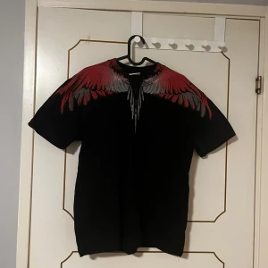 Svart t-shirt med röd- Marcelo Burlon - Säljer en svart t-shirt med korta ärmar och en riktigt snygg vinggrafik i rött och grått över axlar och halsringning, både fram och bak. T-shirten har en klassisk passform och är gjord i mjuk bomull. Perfekt för dig som vill sticka ut med en unik look.