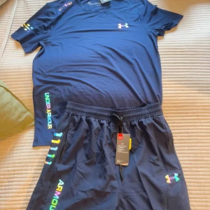Blå träningsshorts från Under Armour M/L. - Snygga marinblå träningsshorts från Under Armour med färgglad logga och text längs benet. Shortsen har elastisk midja med snörning och är gjorda i ett lätt och snabbtorkande syntetmaterial. Perfekta för dig som vill ha en sportig och modern look. M/L.