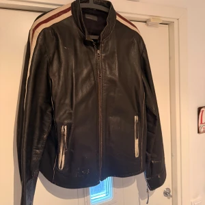 Svart äkta skinnjacka, kort modell - Svart skinnjacka med dragkedja framtill och två fickor med vit-röda detaljer, innerficka, svart silkes foder. Snygga vita och röda ränder längs axlarna och ärmarna ger jackan en sportig vibe. Jackan har ståkrage och är långärmad, perfekt för dig som gillar retrostil och coola detaljer. Använd men fräsch och härlig patina. Inköpt 2005 i Stockholm för 3800kr.