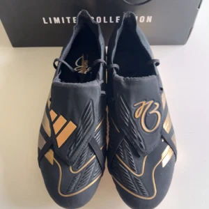 Adidas Predator fotbollsskor limited edition - Snygga Adidas Predator fotbollsskor från Limited Collection i svart med guldiga detaljer och exklusiv guldmetallisk sula. Skorna har en modern, strömlinjeformad design med elastiska band och ikoniska Predator-mönster. Perfekta för dig som vill sticka ut på planen.