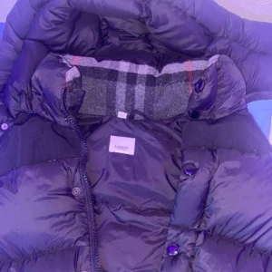 Svart pufferjacka från Burberry - Säljer en svart pufferjacka från Burberry med stor logopatch på ärmen och klassiskt rutigt foder vid kragen. Jackan har dragkedja och knappar framtill, samt en hög krage som ger extra värme. Perfekt för kalla dagar och riktigt snygg streetstil.