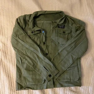 Grön overshirt med fickor - Snygg militärgrön overshirt med fyra stora fickor framtill och svarta knappar. Jackan har klassisk krage och är tillverkad i ett slitstarkt bomullsmaterial. Perfekt för lager-på-lager och chill stil. Långärmad och loose fit.