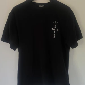 Cactus Jack t-shirt  - Svart t-shirt från Cactus Jack med tryck på bröstet och stor grafisk print på ryggen med texten 'LOOK MOM I CAN FLY' över Travis Scotss mugshot. T-shirten har klassisk passform och är gjord i mjuk bomull. Perfekt för dig som gillar streetwear och Travis Scott vibes.