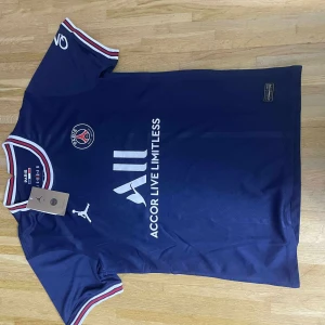 PSG matchtröja blå Puma herr - Snygg blå Paris Saint-Germain fotbollströja från Puma med vita och röda detaljer på krage och ärmslut. PSG-logga och sponsortryck på bröstet, tillverkad i lätt och ventilerande material. Perfekt för dig som älskar fotboll och vill bära klubbens färger.