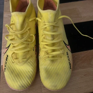 Nike Mercurial gula fotbollsskor - Säljer ett par Nike Mercurial fotbollsskor i en riktigt fräsch gul färg med svarta detaljer och rosa mönster på sidorna. Skorna har snörning, högre skaft och greppig sula för inomhusspel. Perfekta för dig som vill sticka ut på planen.