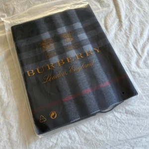 Burberry rutig halsduk i mörkblå ull - Snygg halsduk från Burberry med klassiskt rutigt mönster i mörkblått, grått och röda detaljer. Gjord i mjuk ull och har stilrena fransar i ändarna. Perfekt accessoar för att lyfta din look och hålla dig varm under kyliga dagar.