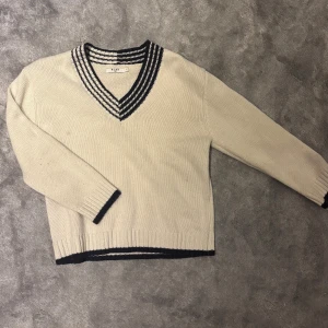 Beige stickad tröja med v-ringning - Snygg beige stickad tröja från NA-KD med v-ringad hals och svarta ränder runt kragen, ärmsluten och nederkanten. Tröjan är långärmad och gjort på ett lite tyngre garn, mysig att ha både hemma och ute. Finns tyvärr en liten fläck på sig. Kan säkert gå bort i kemtvätt.