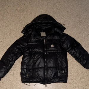Svart Moncler dunjacka med huva - Snygg svart dunjacka från Moncler med glansig finish, huva och dragkedja framtill. Jackan har två fickor med dragkedja och en bröstficka med Moncler-logga. Perfekt för kalla dagar och riktigt stilren look. Moncler jacka – svart och varm Snygg vinterjacka från Moncler i svart färg. Väldigt varm och bekväm, perfekt för kalla dagar. Jackan är i fint skick och ser bra ut. Obs: Höger ficka är lite sprucken men det går lätt att laga hos en skräddare.!!