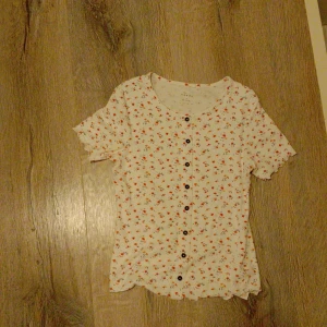 Blommig topp från basic needs - Supersöt blommig topp från basic needs med små röda och rosa blommor på vit botten. Toppen har korta ärmar, rund halsringning och svarta knappar framtill. Materialet är ribbat och stretchigt, perfekt för en chill och söt look. Priset går att diskuteras <333