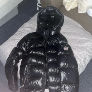 Svart Colmar pufferjacka med huva - Säljer en svart, glansig pufferjacka från Colmar med stor huva och dragkedja framtill. Jackan har tydliga quiltade partier och logga på ärmen. Insidan är fodrad med Colmar-tryck. Perfekt för kalla dagar och riktigt snygg streetstil.