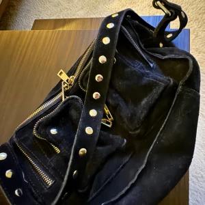 Svart mocka axelväska med nitar -  svart axelväska i mocka med silvtiga dragkedjor och stora silverfärgade nitar på axelremmen. Från märket NUNOO.