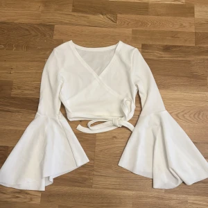 Vit omlottopp med utsvängda ärmar - Säljer en helt oanvänd vit omlottopp med långa, vida trumpetärmar och knytband i midjan. Toppen är croppad och har en snygg v-ringning framtill. Perfekt för dig som vill sticka ut med en trendig och stilren look.