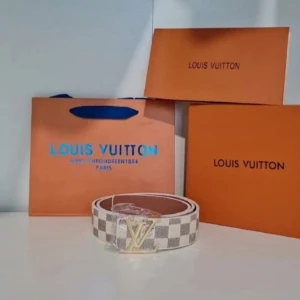 rutigt bälte i skinn - Snyggt bälte från  med klassiskt rutigt mönster i vitt och grått. Bältet har en ikonisk spänne i guld och är tillverkat i skinn. Perfekt accessoar för att lyfta din outfit med lyxig känsla.