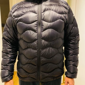 Svart dunjacka Peak Performance XL - Säljer en svart dunjacka från Peak Performance i storlek XL. Jackan har vågformat quiltat mönster, dragkedja framtill och två sidofickor med dragkedja. Perfekt för kalla dagar och riktigt snygg passform. Passar även en L. Jackan är som ny! 
