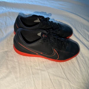 Svarta Nike fotbollsskor med röd sula - Säljer ett par svarta Nike fotbollsskor med snygga röda detaljer och röd sula. Skorna har snörning och är tillverkade i syntetmaterial för en lätt känsla på planen. Perfekta för dig som vill sticka ut med stilrena och sportiga skor.