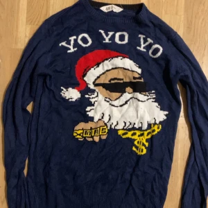 Blå jultröja med cool tomte från H&M - Mörkblå stickad jultröja från H&M med en rolig tomte i solglasögon, guldkedja och texten 'YO YO YO' framtill. Tröjan har långa ärmar och ribbade muddar. Perfekt för att sticka ut på julfirandet!