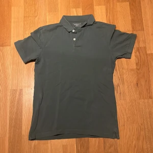 Grön pikétröja från Dressmann - Säljer en stilren grön pikétröja från Dressmann i storlek S. Tröjan har klassisk krage, korta ärmar och knäppning med två knappar framtill. Perfekt för en clean och enkel look. Tillverkad i mjuk bomull som känns skön mot huden.