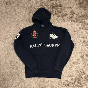 Mörkblå hoodie Polo Ralph Lauren - Mörkblå hoodie från Polo Ralph Lauren i junior storlek XL, vilket motsvarar runt S-M. Riktigt snygg hoodie med stor vit logga och text framtill, broderad patch på bröstet och siffra 3 på ryggen. Finns lite tecken på användning. Skriv vid vidare funderingar!
