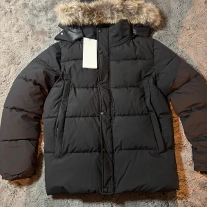 Svart dunjacka med päls från Canada Goose - Canada Goose dunjacka i svart med fluffig päls på huvan och klassisk logga på ärmen. Jackan har långa ärmar, dragkedja och knappar framtill samt två stora fickor. Perfekt för kalla vinterdagar och har en riktigt schysst oversized look.