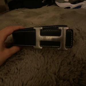 Svart H bälte med H-spänne - Säljer ett svart bälte från H med ikoniskt H-format metallspänne. Bältet har vita kontrastsömmar längs kanterna och är tillverkat i skinn. Perfekt accessoar för att lyfta vilken outfit som helst.