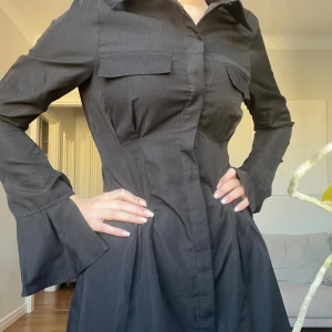 Svart skjortklänning  - Jättefin kort, svart skjortklänning från pretty little thing. Klänningen har en figurnära passform kring midjan med en lösare passform vid kjolen. Den har också knytning vid ärmarna och väldigt fina rätt så breda manschetter vid handlederna🩷 köptes för 599kr, aldrig använd🩷 