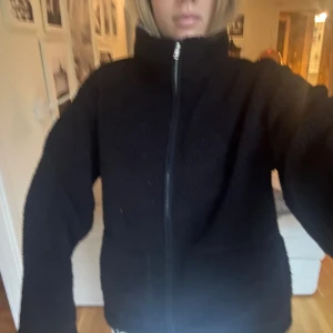 Svart teddyjacka med dragkedja - Mysig svart teddyjacka med hög krage och hel dragkedja framtill. Jackan har en loose passform och är perfekt för kyliga dagar. Tillverkad i mjukt fleece-material som ger en skön och avslappnad vibe.