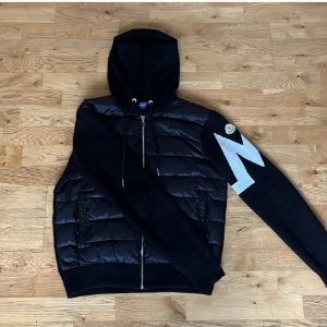 Svart pufferjacka från Moncler med huva - Svart pufferjacka från Moncler med huva och dragkedja. Jackan har quiltad framsida i glansig polyester och stickade ärmar. På ena ärmen finns en stor vit Moncler-logga. Perfekt för dig som vill ha en sportig och stilren look.