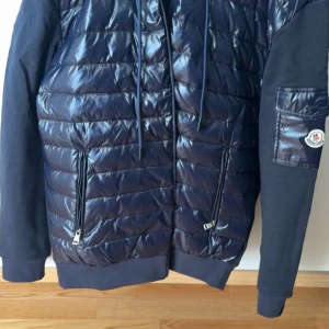 Mörkblå pufferjacka från Moncler - Snygg mörkblå pufferjacka från Moncler med glansig quiltad framsida och ärmficka med logotyp. Jackan har dragkedjor framtill och på fickorna samt ribbade muddar.  Med NFC, passar M också.  BUDA