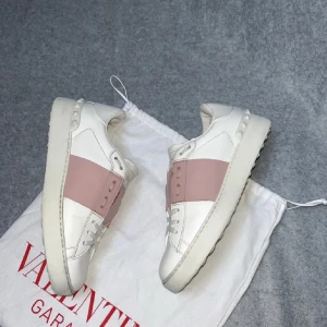 Valentino Open sneakers vit/rosa - Snygga Valentino Open sneakers i vitt skinn med bred rosa panel över mitten. Klassisk rund tå och platt sula, med coola nitar på hälen. Perfekt för dig som gillar exklusiva märken och stilren design.