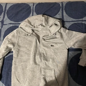 Grå hoodie från Lacoste med dragkedja - Säljer en grå hoodie från Lacoste i regular fit med klassisk krokodillogga på bröstet. Tröjan har huva med snörning, dragkedja framtill och kängurufickor. Perfekt för chill dagar och enkel att matcha med jeans eller joggers.