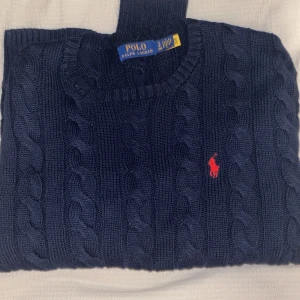 Grå kabelstickad tröja Polo Ralph Lauren - Säljer en klassisk grå kabelstickad tröja från Polo Ralph Lauren med röd broderad logga på bröstet. Tröjan är stickad i mjuk bomull och har rund halsringning samt långa ärmar. Perfekt för dig som gillar stilren och tidlös design.