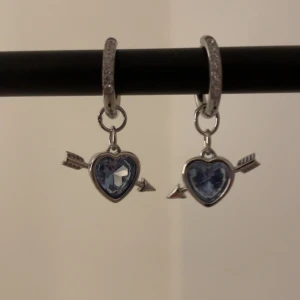 Silverörhängen med blå hjärtan och pil - Säljer ett par snygga örhängen i silverfärg med glittrande blå hjärtformade hängen och pil-detaljer. Ringarna har små gnistrande stenar och hängena är formade som hjärtan med en pil igenom, vilket ger en cool och romantisk vibe.