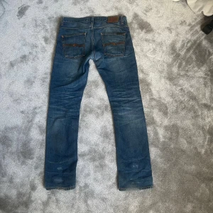 Blå jeans från Nudie Jeans, slim/straight fit - Säljer ett par klassiska blå jeans från Nudie Jeans med snyggt slitna detaljer och kontrastsömmar. Modellen har raka ben och normal passform, tillverkade i bomull. Perfekta för dig som gillar en avslappnad och tidlös stil. Storlek W 31 L 32