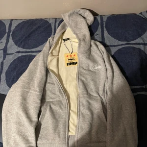 Grå hoodie från Cryptz med dragkedja - Säljer en stilren grå hoodie från  corteiz med broderad logga på bröstet. Tröjan har huva, dragkedja framtill och två fickor. Perfekt för dig som gillar en clean och avslappnad look. Materialet är mjukt och skönt, passar till många outfits.