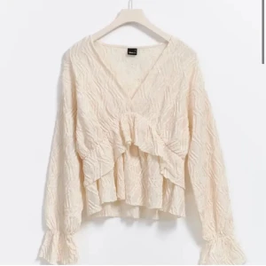Beige omlottblus med volang från Gina Tricot - Säljer en beige omlottblus från Gina Tricot med v-ringning och volangdetalj framtill. Blusen har långärmade ärmar med lätt rynk vid mudden och ett snyggt, krinklat tyg som ger en cool struktur. Perfekt att styla med jeans eller kjol för en trendig look.