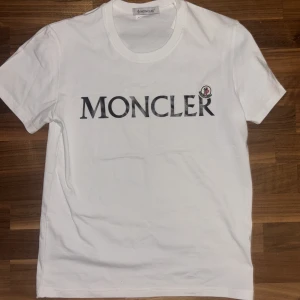Vit Moncler t-shirt - Vit t-shirt från Moncler med stor svart logotyp framtill och klassisk rund hals. T-shirten har korta ärmar och ett litet Moncler-märke på bröstet. Tillverkad i mjuk bomull för en clean och stilren look. Passar mig som är 184cm. Helt ny! 