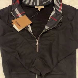 Svart Burberry vindjacka med rutig huva - Svart vindjacka från Burberry med klassisk rutig huva i grått, vitt och rött. Jackan har dragkedja framtill, ribbade muddar och broderad Burberry-logga på bröstet. Perfekt för dig som gillar stilrena och ikoniska detaljer. Den är S men passar XS oxå 