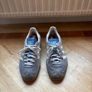 Adidas Samba grå mocka sneakers - Adidas Samba sneakers i grå mocka med vita klassiska ränder på sidorna och blå plös med Adidas-logga. Skorna har snörning och en gummisula med mönster för extra grepp. Perfekt för dig som gillar retrostil och sportig vibe.