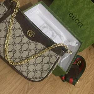 Gucci axelväska med kedja - Snygg axelväska från Gucci i klassisk GG Supreme-mönster. Beige och brunt tyg med bruna skinn-detaljer, guldfärgad kedja och GG-logo framtill. Väskan har dragkedja och medföljande grön-röd axelrem. Perfekt för dig som vill ha en ikonisk designerlook🩷