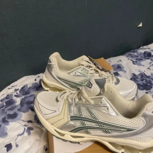 Vita ASICS sneakers med silverdetaljer - Säljer ett par fräscha ASICS sneakers i vit och silver med coola meshpaneler och Ortholite innersula för extra komfort. Skorna har snörning och en chunky platt sula som ger en sportig vibe. Perfekta för dig som gillar stilrena och moderna träningsskor.