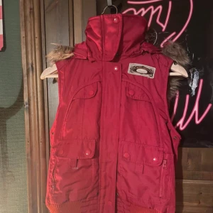 Röd dunväst från Vero Moda - Röd dunväst från Vero Moda med hög krage, avtagbar huva med fuskpäls och flera fickor framtill. Västen har ribbade detaljer nedtill och coolt Arctic Explorer-märke på bröstet. Perfekt för lager-på-lager under kyliga dagar.